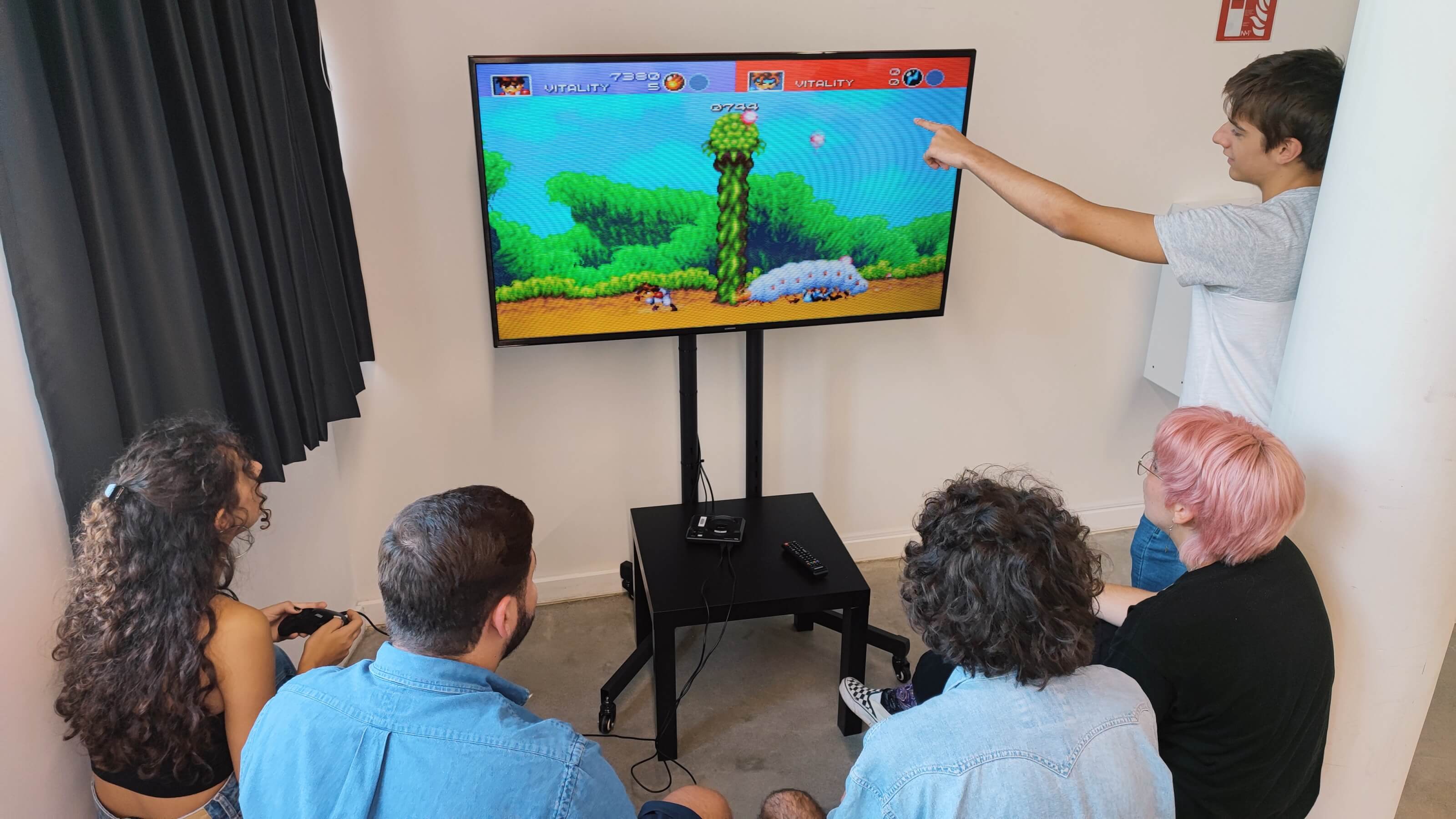 A group of 5 students play a Sega Mega Drive game around a tv.