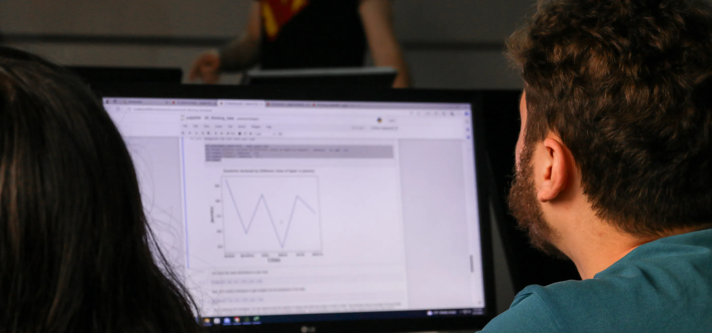 A student looking to the front of the class, while his screen shows the graphs resulting from his code