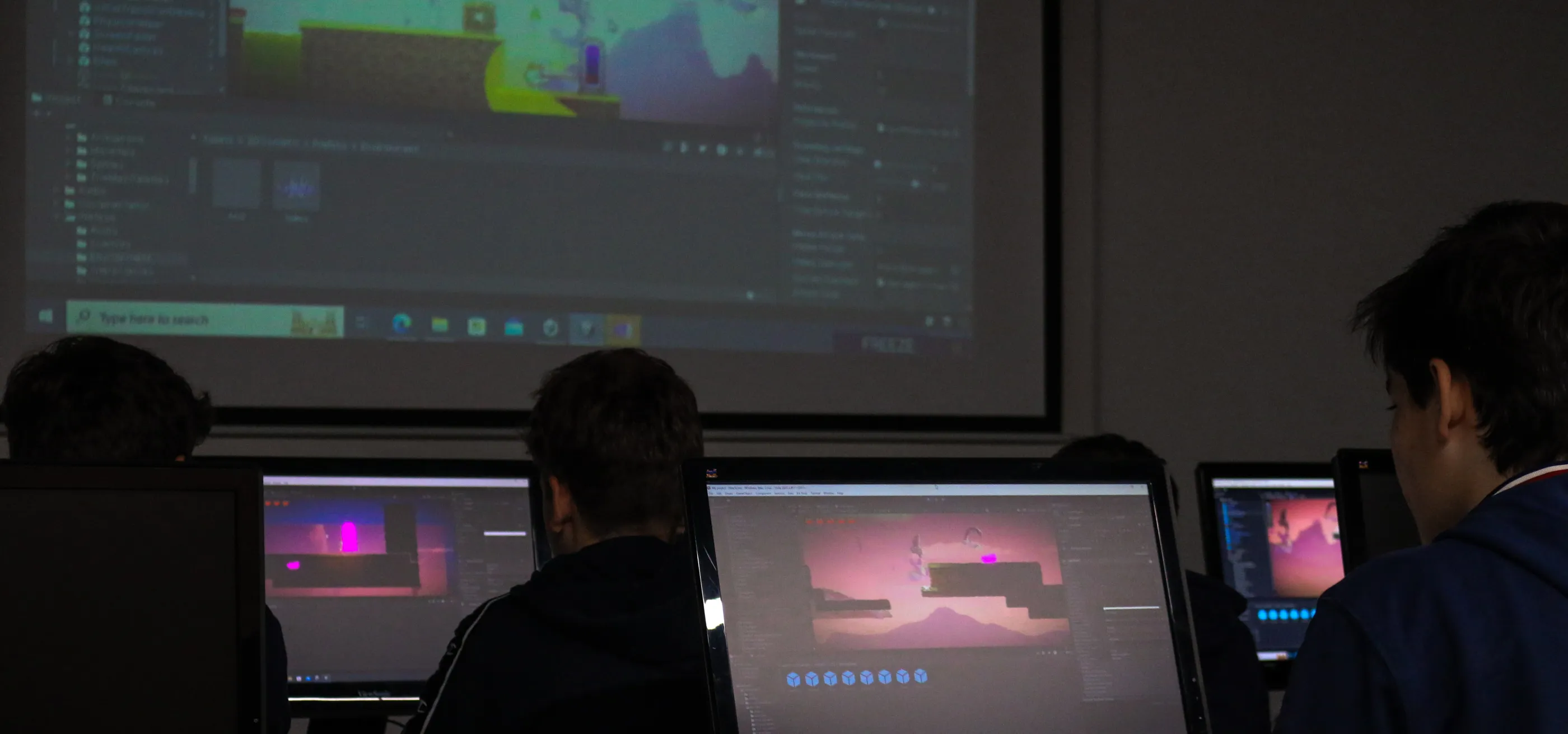 Estudiantes trabajan en un proyecto utilizando el motor Unity mientras el instructor muestra algo en la pantalla grande en el fondo.