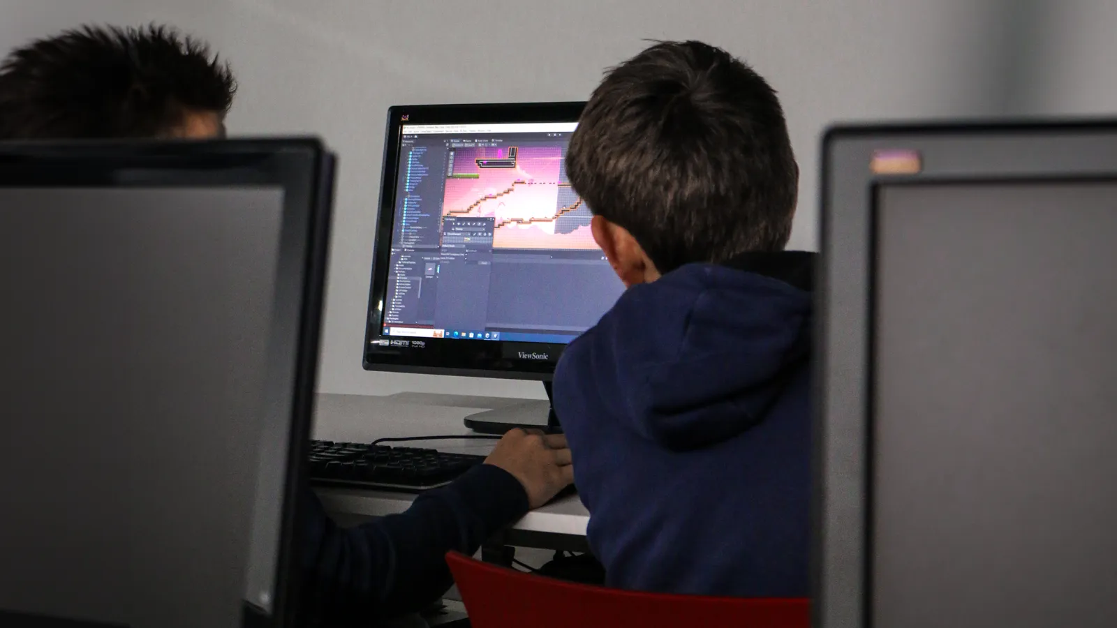 Entre las pantallas, dos estudiantes trabajan juntos en la creación de su proyecto de juego. La pantalla muestra un videojuego con plataformas.
