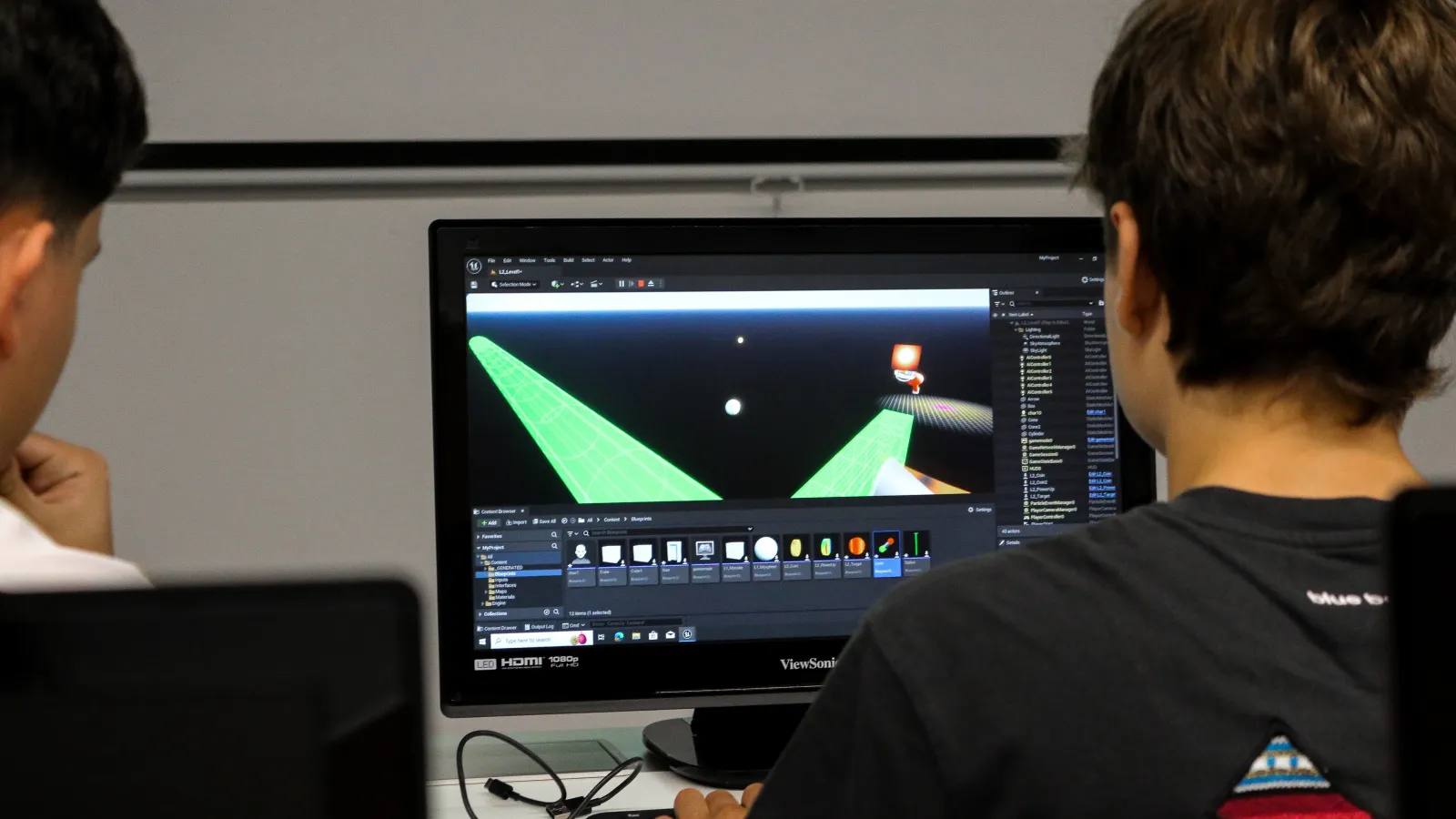 Dos estudiantes que asisten al taller trabajan en la misma computadora usando el editor de Unreal Engine. El proyecto muestra un escenario oscuro y laberíntico.