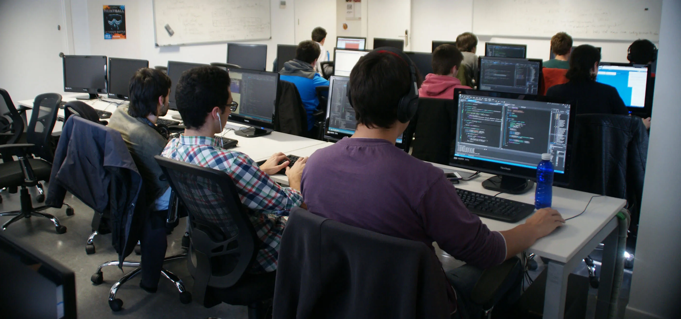 Multiple students sit in at computer workstations while working on programming