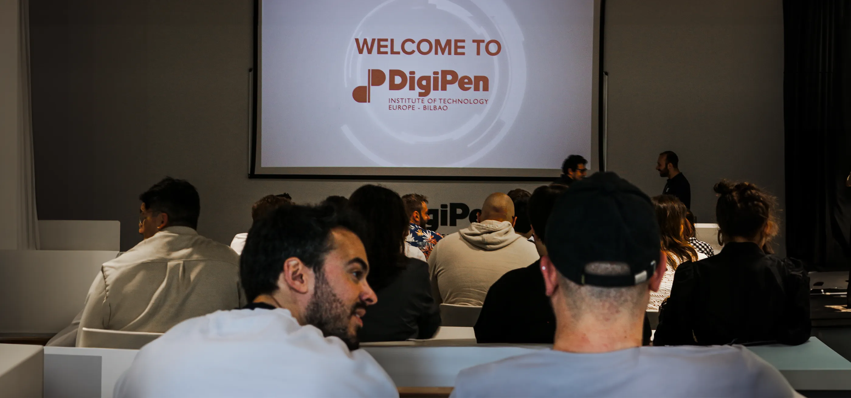 En el interior de un aula de DigiPen Europe - Bilbao, alumnos y profesores sentados y esperando el comienzo de una presentación, con una gran pantalla en la que se muestra el logotipo de DigiPen y el mensaje «Bienvenido a DigiPen».