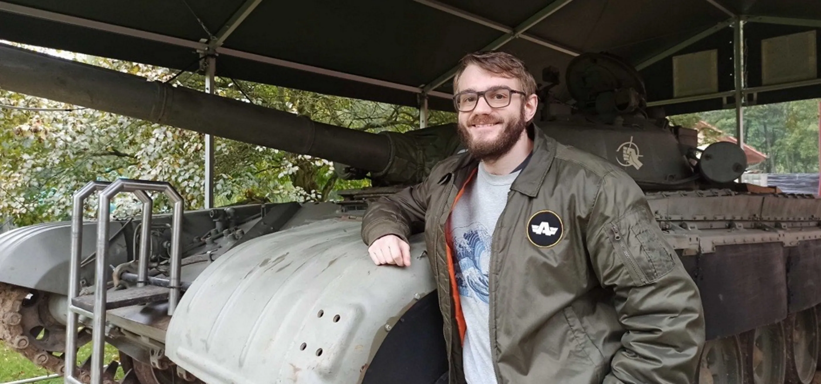 Jokin Bilbao smiling and standing next to an old military tank, wearing a Bohemia Interactive jacket during a visit to a military exhibition.