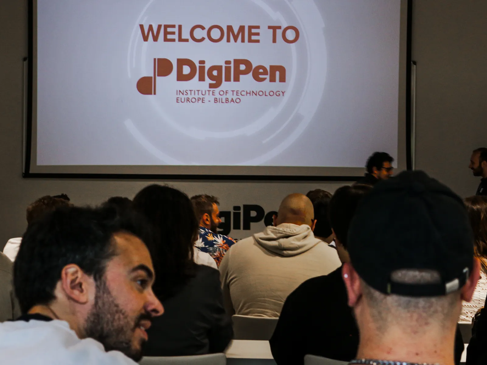 En el interior de un aula de DigiPen Europe - Bilbao, alumnos y profesores sentados y esperando el comienzo de una presentación, con una gran pantalla en la que se muestra el logotipo de DigiPen y el mensaje «Bienvenido a DigiPen».