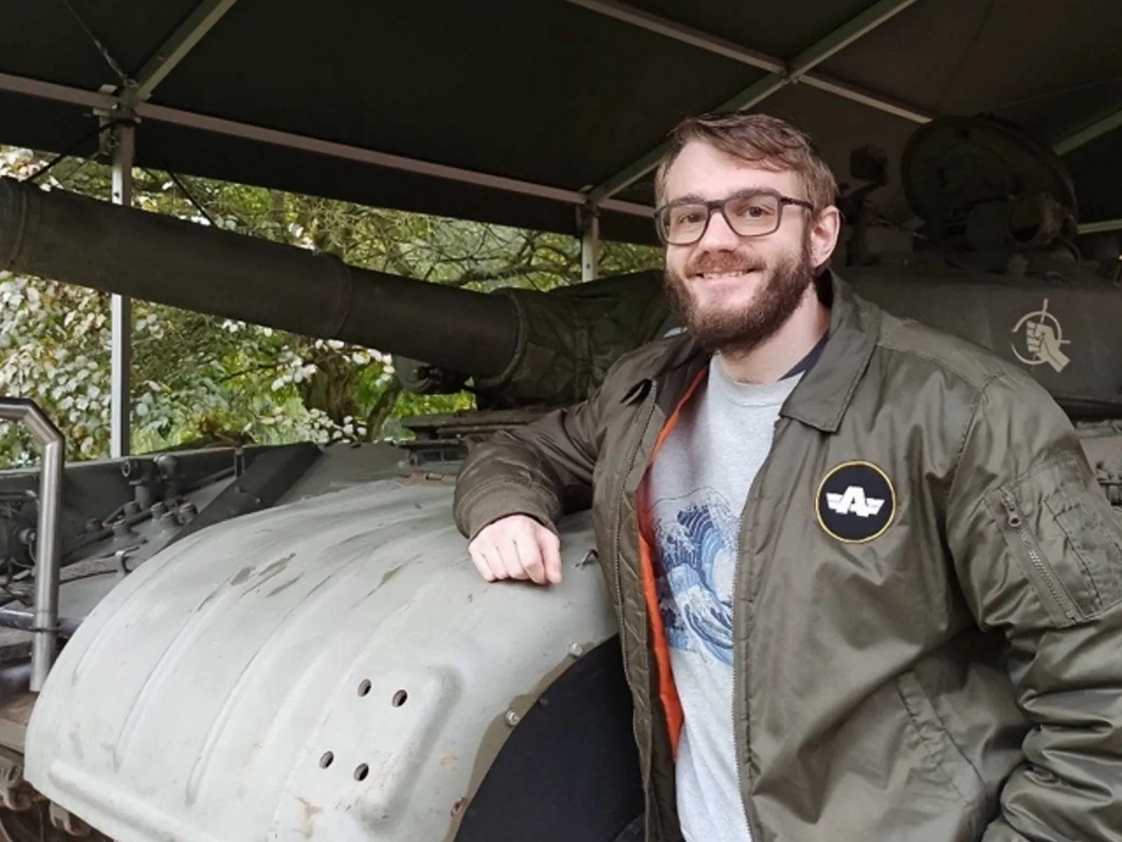 Jokin Bilbao smiling and standing next to an old military tank, wearing a Bohemia Interactive jacket during a visit to a military exhibition.