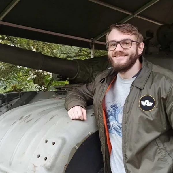 Jokin Bilbao smiling and standing next to an old military tank, wearing a Bohemia Interactive jacket during a visit to a military exhibition.