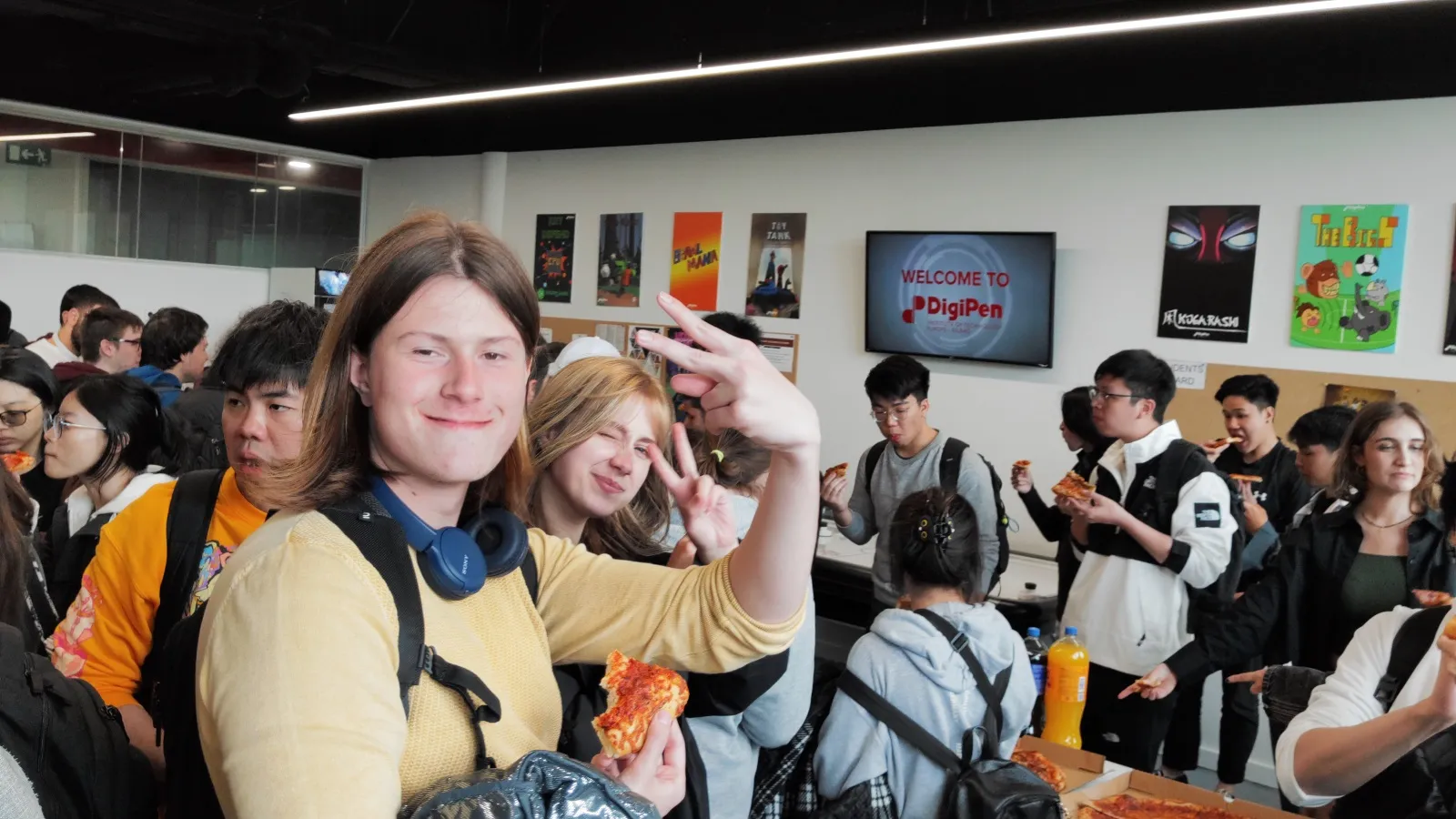 Un grupo de estudiantes en DigiPen Europe-Bilbao sonriendo y comiendo pizza durante un evento social, con obras de arte y una pantalla de bienvenida visibles al fondo.