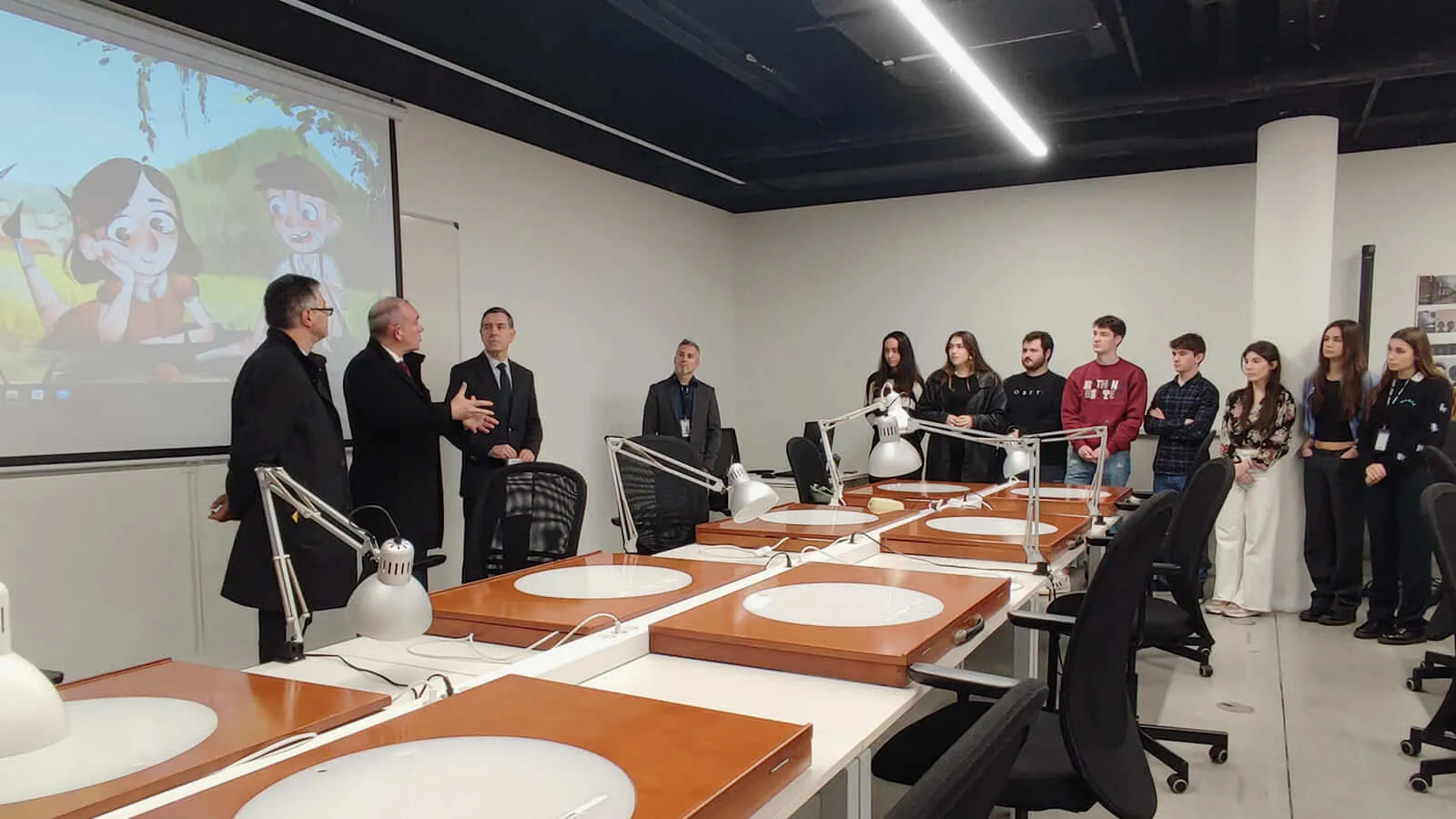 A group of students stand in a classroom listening to 3 men talk.