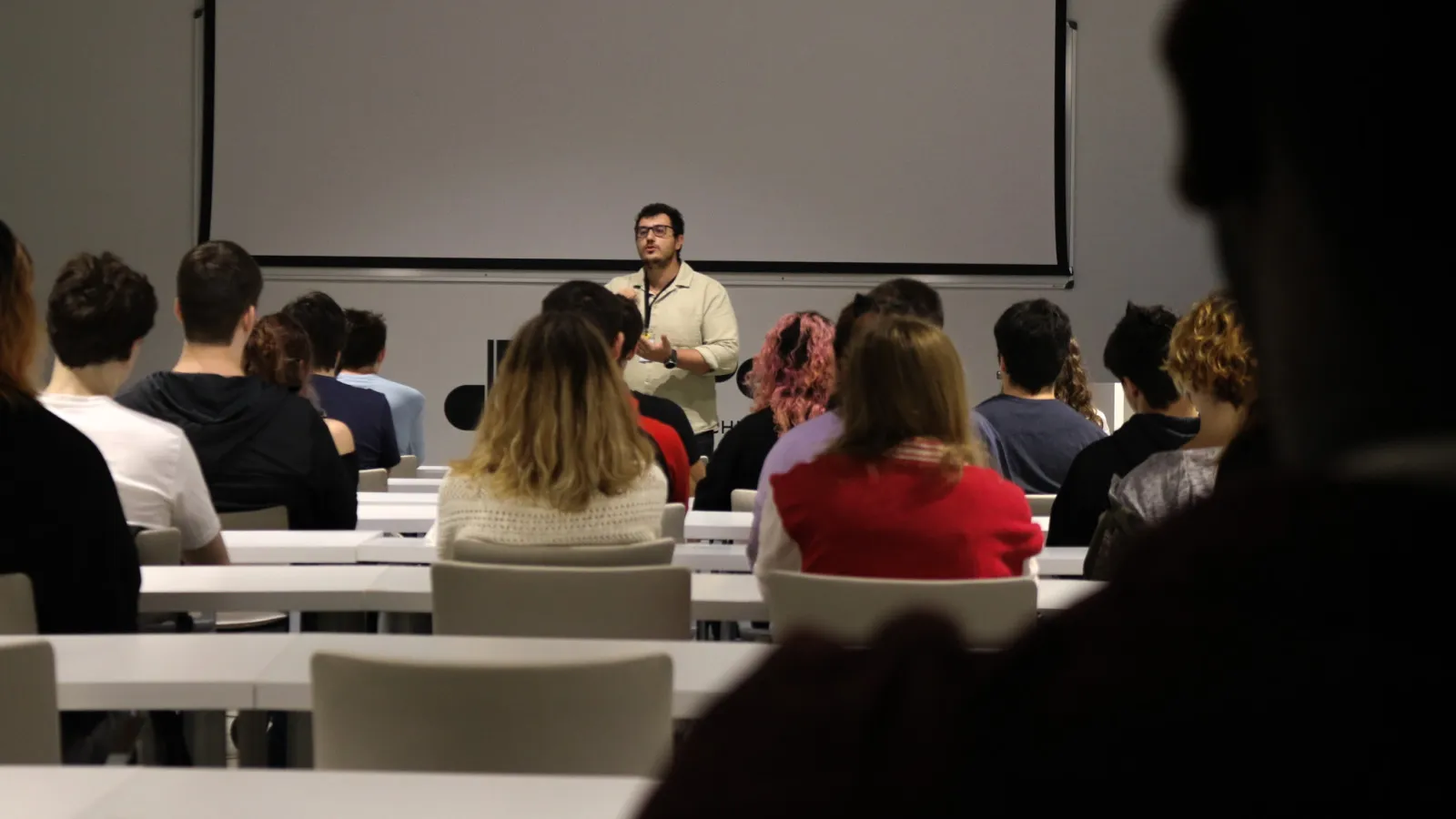 Un profesor presenta a una clase de estudiantes sentados de frente a él, con sus espaldas hacia la cámara.