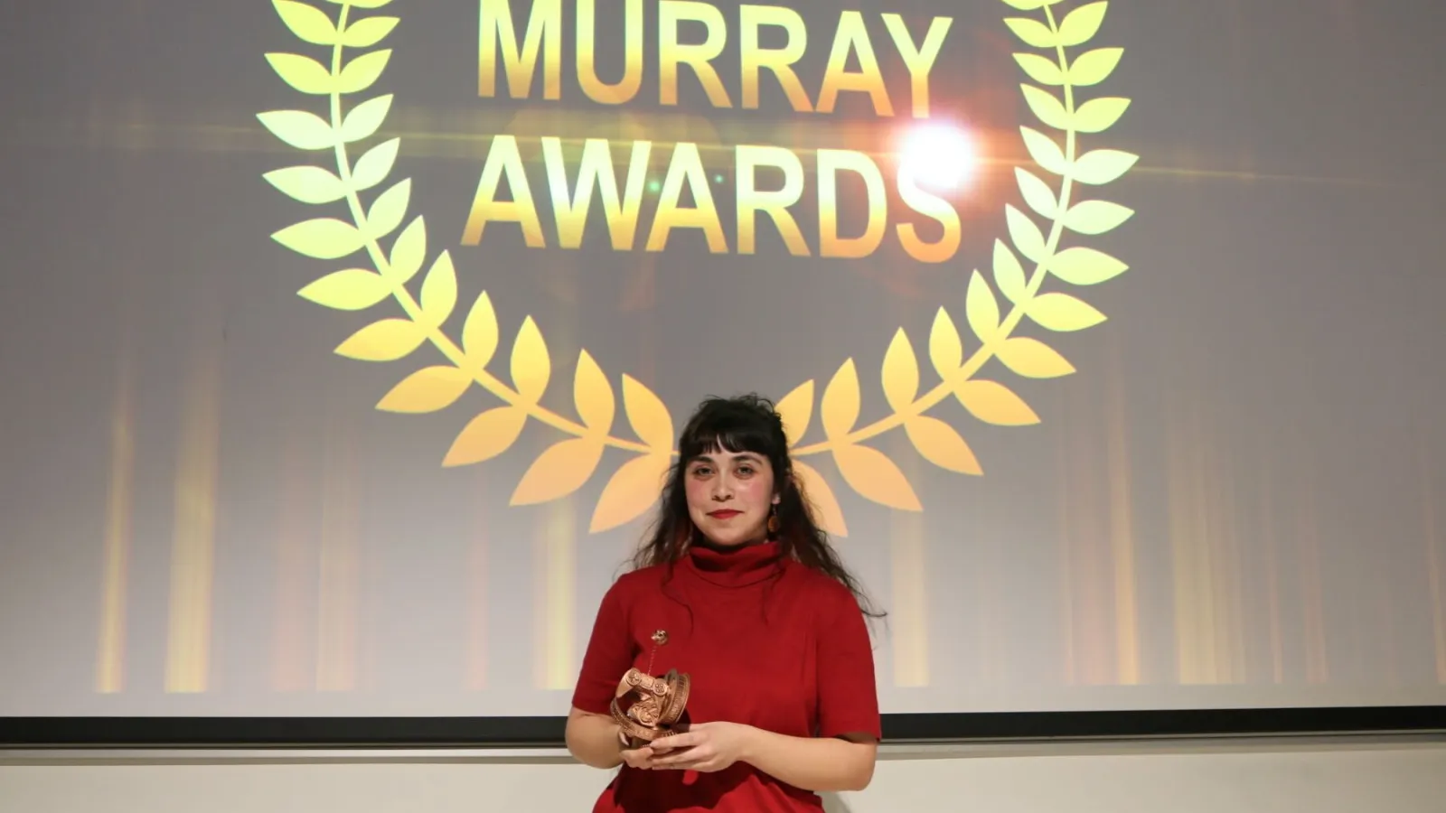 A student holds her trophy and smiles as she stands in front of the awards show banner.