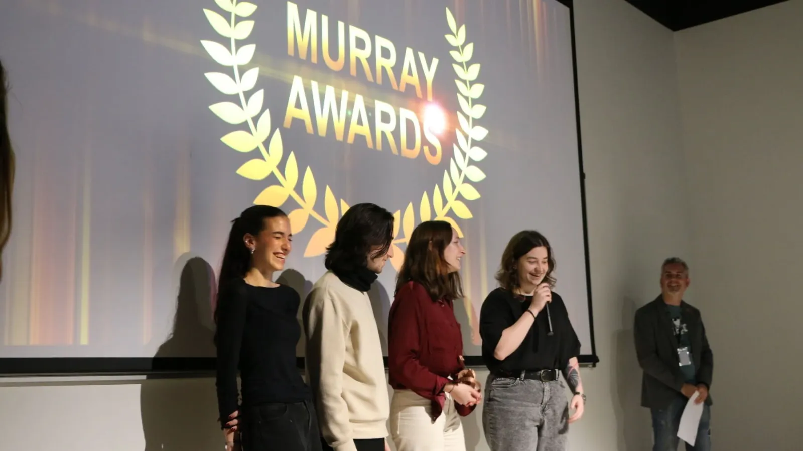 The presenter of the awards show steps aside to allow a student film team a moment to speak after winning an award.