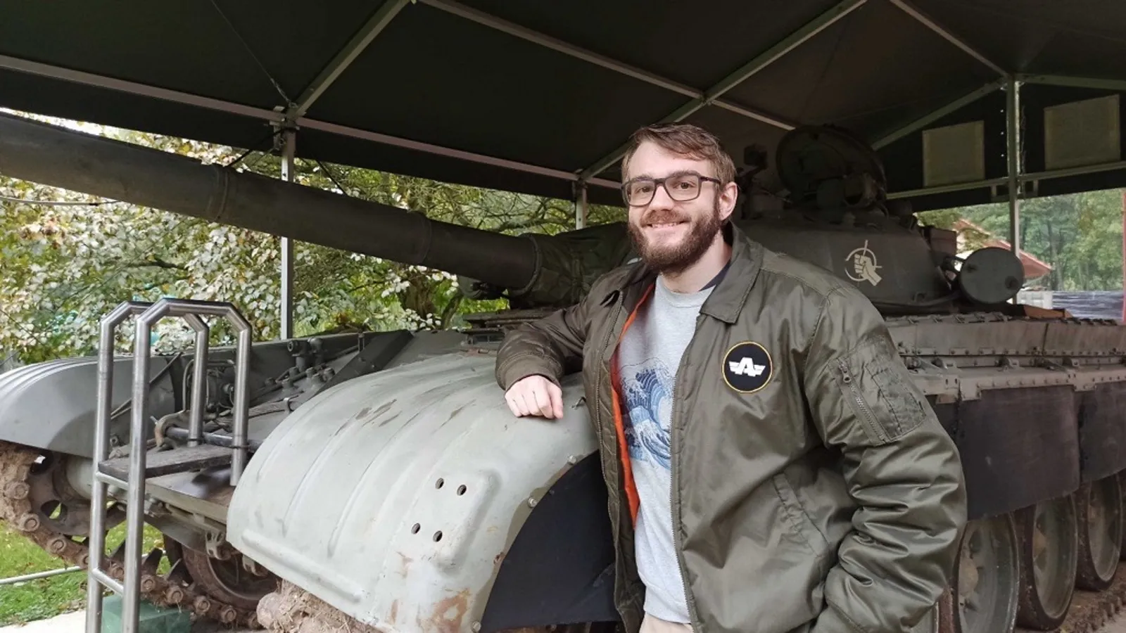 Jokin Bilbao smiling and standing next to an old military tank, wearing a Bohemia Interactive jacket during a visit to a military exhibition.