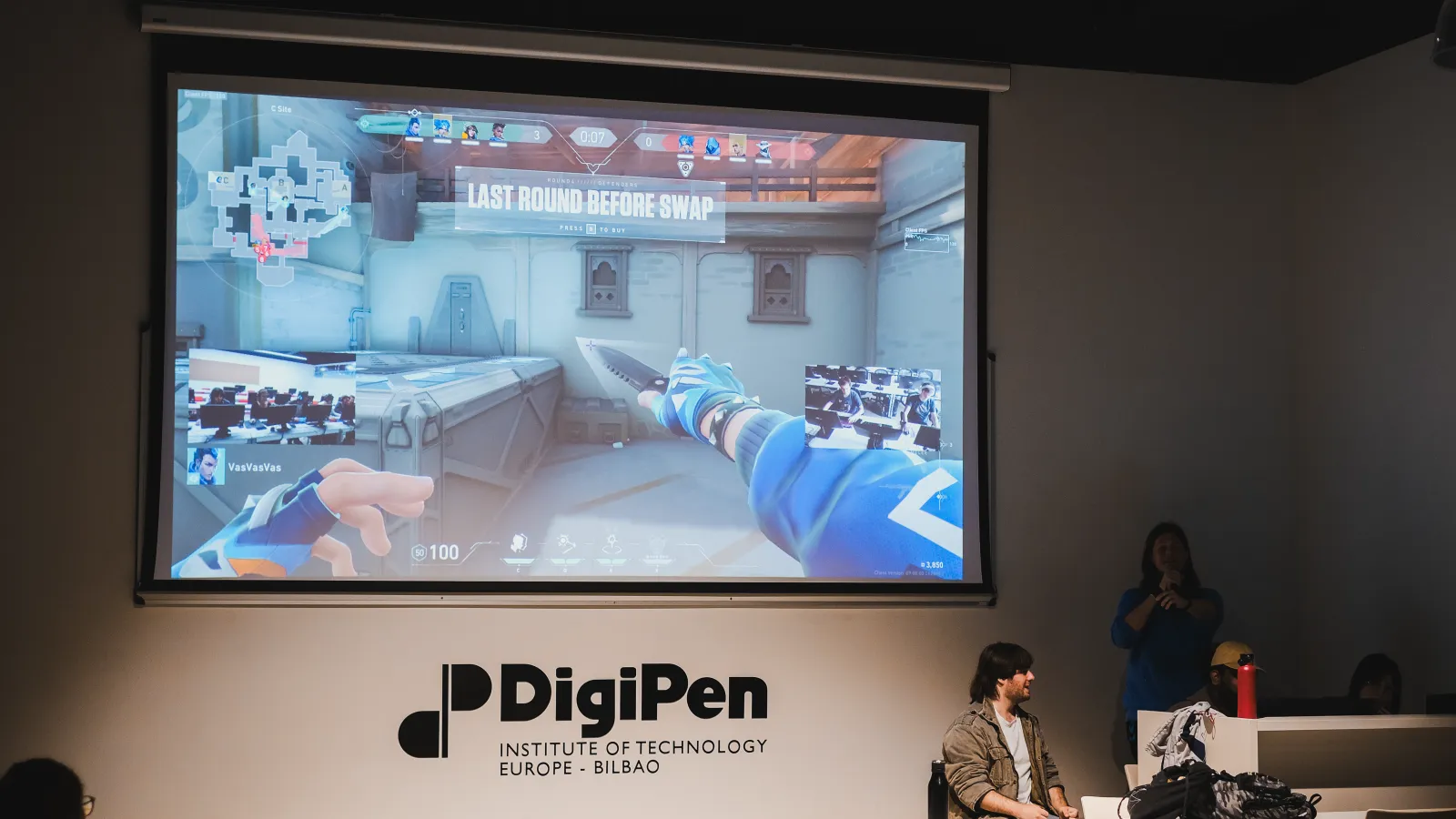 A large screen at DigiPen Europe-Bilbao showing a first-person perspective of a Valorant match, with students playing the game in a classroom setting visible in an inset