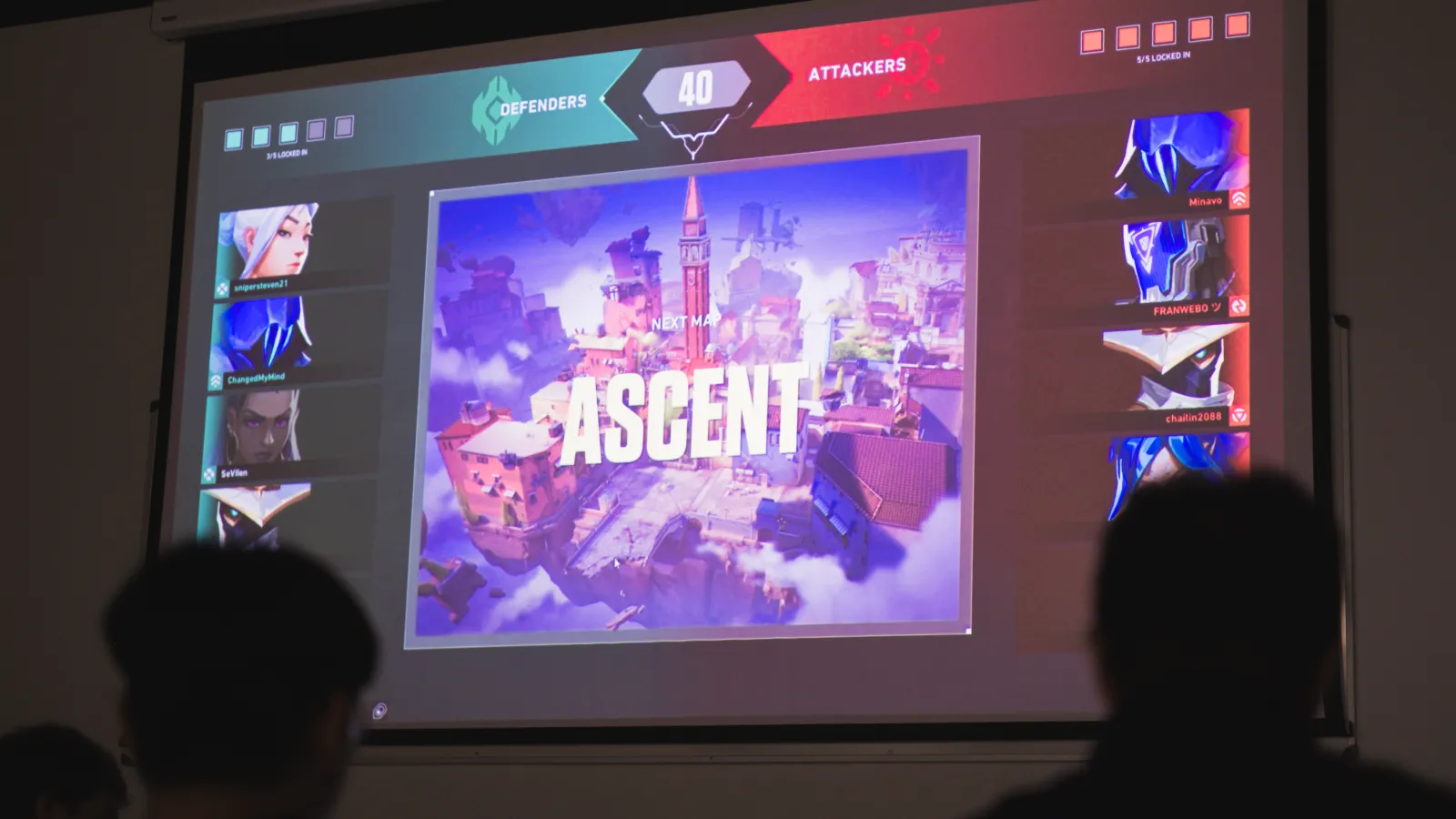 Pantalla grande en DigiPen Europe-Bilbao que muestra la pantalla de selección del mapa «Ascenso» en una partida de Valorant, con siluetas de estudiantes observando en primer plano.