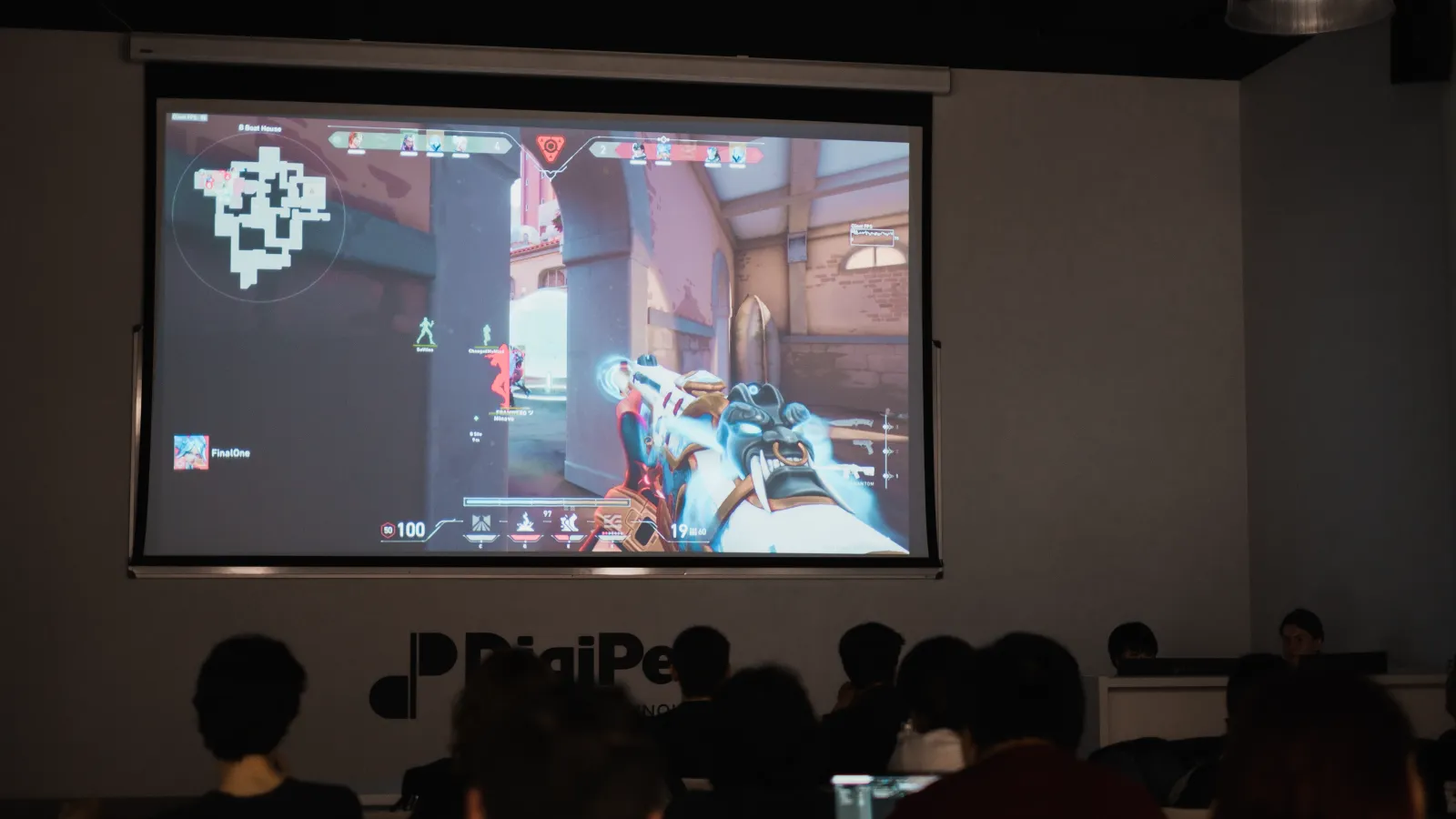 A large screen at DigiPen Europe-Bilbao displaying a Valorant match with the game map and player positions, while students watch attentively.