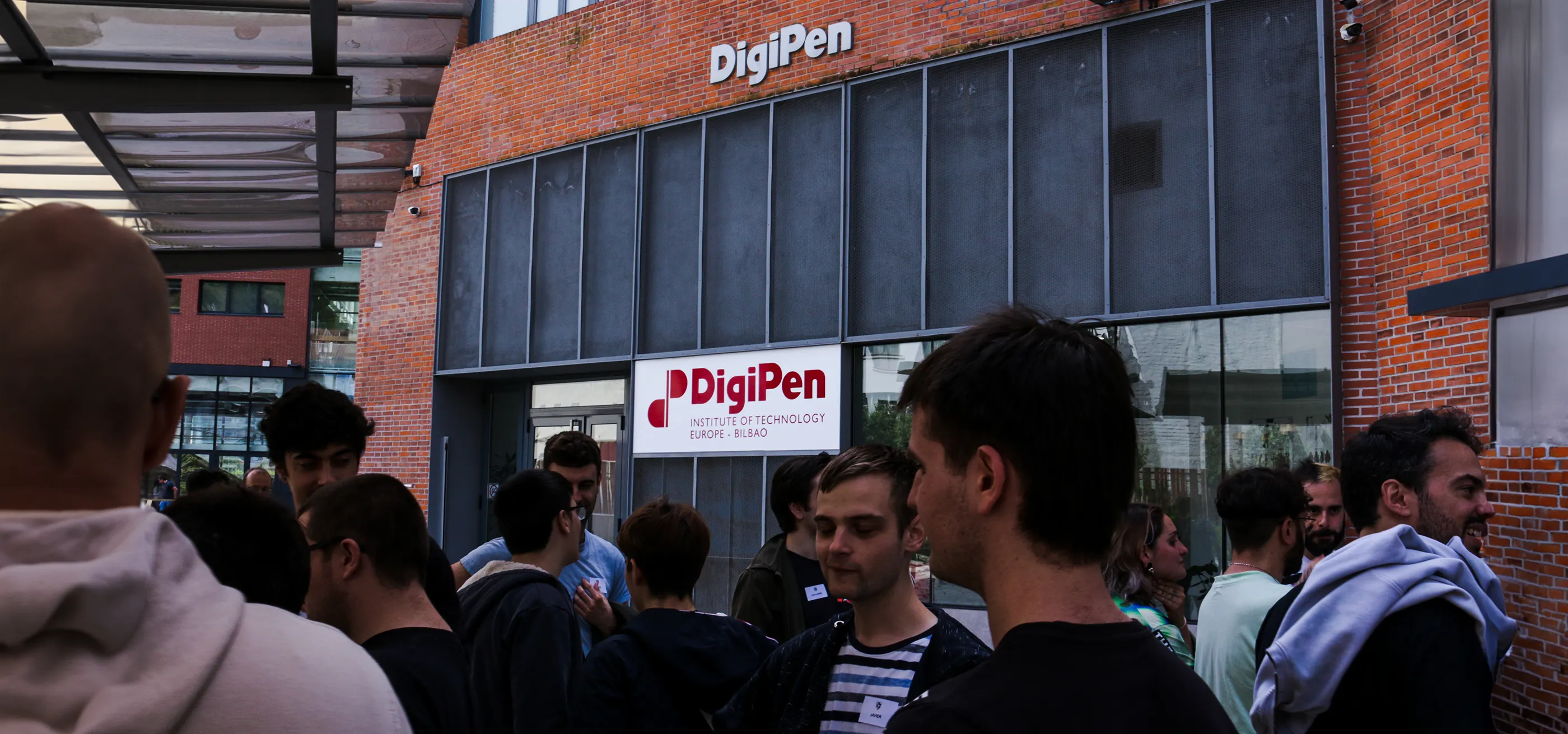 Un grupo de estudiantes reunidos en el exterior del Instituto Tecnológico DigiPen Europa - Bilbao, entablando conversaciones cerca de la entrada del campus, con el logotipo de DigiPen visible al fondo.